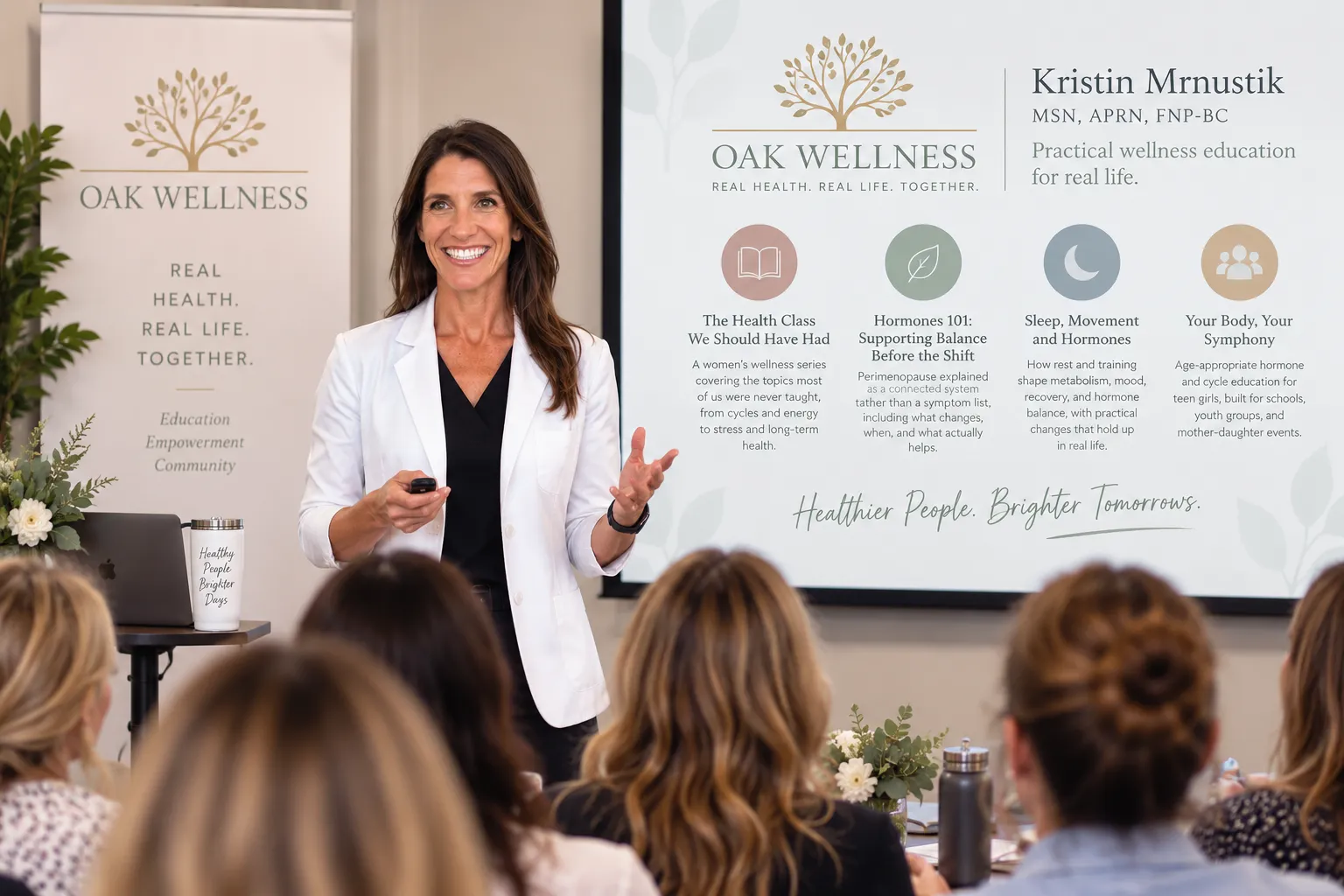 Kristin speaking to a seated audience beside an Oak Wellness banner and slide